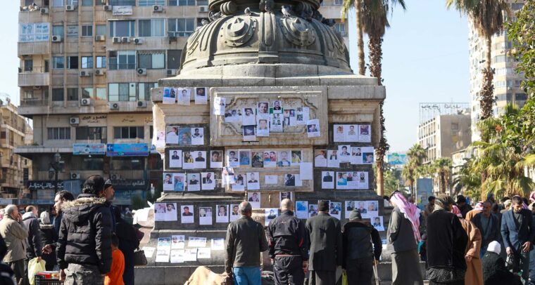 Personas reunidas alrededor de una estructura donde cuelgan fotografías de familiares desaparecidos, en una plaza urbana en Alepo (Siria). Se observan edificios, palmeras y varias personas mirando las imágenes y oraciones expuestas. HTS