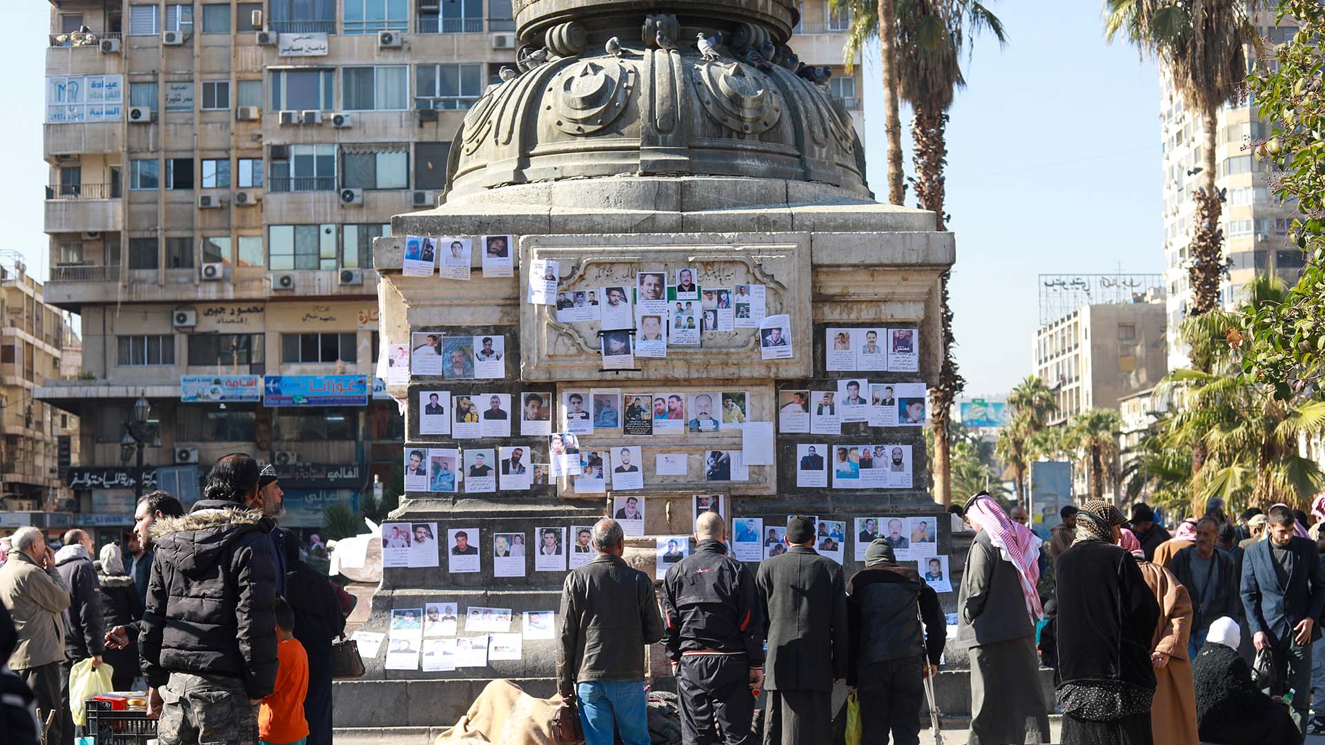 Personas reunidas alrededor de una estructura donde cuelgan fotografías de familiares desaparecidos, en una plaza urbana en Alepo (Siria). Se observan edificios, palmeras y varias personas mirando las imágenes y oraciones expuestas. HTS