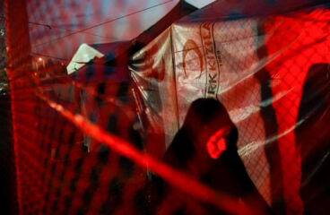 Una mujer vestida de negro avanza por un campo de personas refugiadas durante la noche. La escena está iluminada por una luz roja que proviene del exterior de la malla metálica. Al fondo, se aprecian tiendas de campaña cubiertas con lonas. refugiados