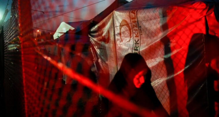 Una mujer vestida de negro avanza por un campo de personas refugiadas durante la noche. La escena está iluminada por una luz roja que proviene del exterior de la malla metálica. Al fondo, se aprecian tiendas de campaña cubiertas con lonas. refugiados