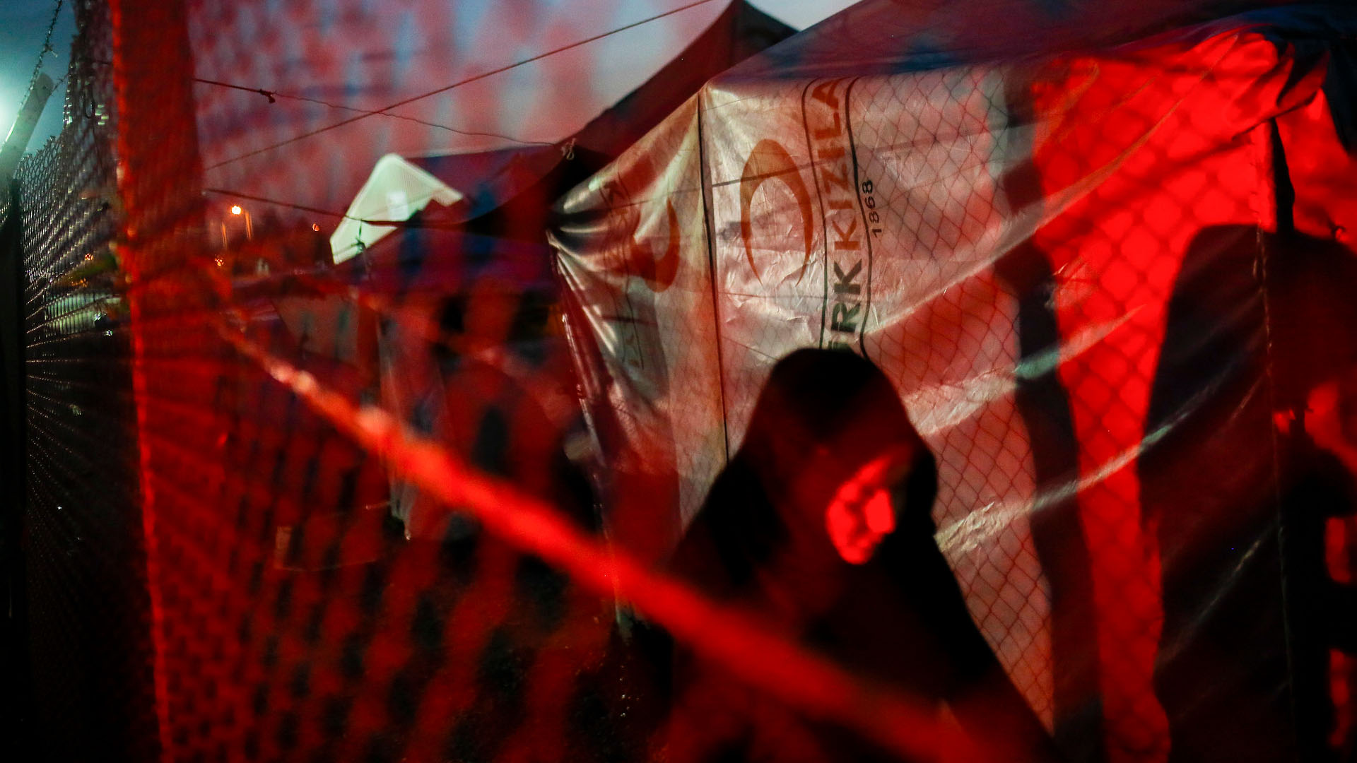 Una mujer vestida de negro avanza por un campo de personas refugiadas durante la noche. La escena está iluminada por una luz roja que proviene del exterior de la malla metálica. Al fondo, se aprecian tiendas de campaña cubiertas con lonas. refugiados