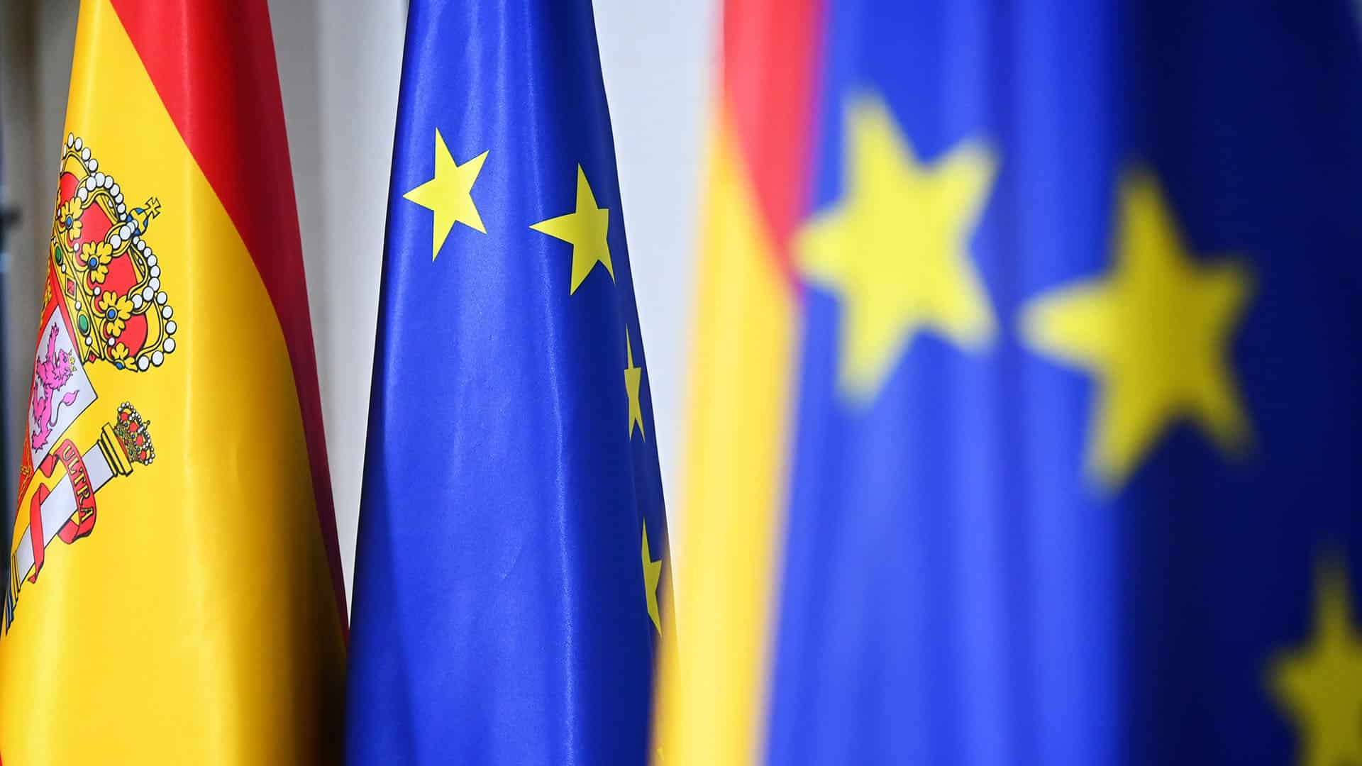 Banderas de España y de la Unión Europea en un acto oficial. La bandera de España, con franjas rojas y amarillas y su escudo nacional, junto a la bandera de la Unión Europea, de color azul con estrellas amarillas. Parlamento Europeo