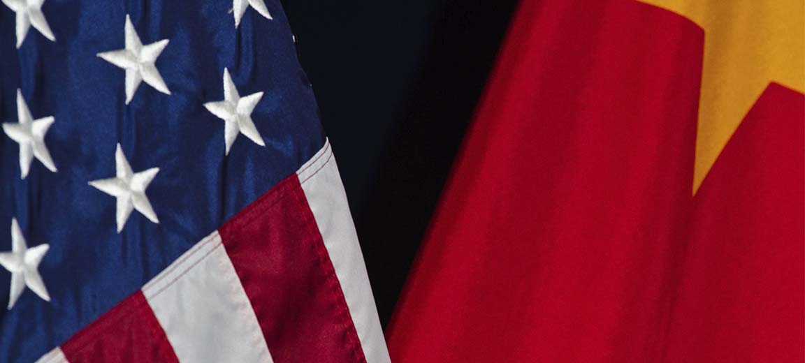 Primer plano de las banderas de Estados Unidos y China, parcialmente visibles sobre un fondo oscuro. La bandera de Estados Unidos muestra estrellas blancas sobre azul y franjas rojas y blancas, mientras la de China presenta fondo rojo con una estrella amarilla. Geopolítica