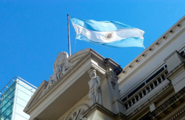 Vista desde abajo de la fachada del edificio del Banco Central de la República Argentina con una bandera argentina azul celeste y blanca ondeando bajo un cielo despejado. Plan de estabilización