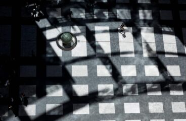 Vista cenital de una plaza con un mosaico de baldosas blancas y negras. Las sombras proyectadas por una estructura forman patrones geométricos sobre el pavimento. Varias personas transitan por el lugar, mientras una esfera de piedra reposa sobre un pedestal circular. Orden liberal