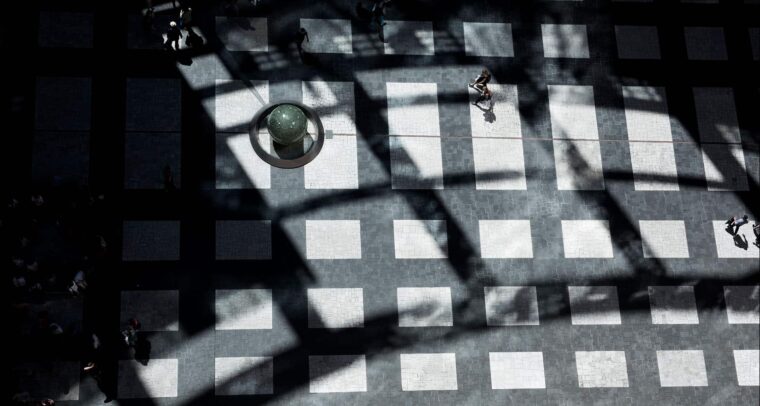 Vista cenital de una plaza con un mosaico de baldosas blancas y negras. Las sombras proyectadas por una estructura forman patrones geométricos sobre el pavimento. Varias personas transitan por el lugar, mientras una esfera de piedra reposa sobre un pedestal circular. Orden liberal