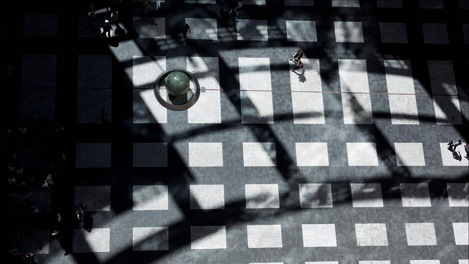 Vista cenital de una plaza con un mosaico de baldosas blancas y negras. Las sombras proyectadas por una estructura forman patrones geométricos sobre el pavimento. Varias personas transitan por el lugar, mientras una esfera de piedra reposa sobre un pedestal circular. Orden liberal