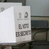 Persona votando detrás de un biombo blanco con la inscripción "El voto es secreto" y logotipos del CNE y la República del Ecuador, en un aula adaptada como recinto electoral con escritorios visibles al fondo. Elecciones