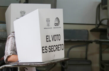 Persona votando detrás de un biombo blanco con la inscripción "El voto es secreto" y logotipos del CNE y la República del Ecuador, en un aula adaptada como recinto electoral con escritorios visibles al fondo. Elecciones