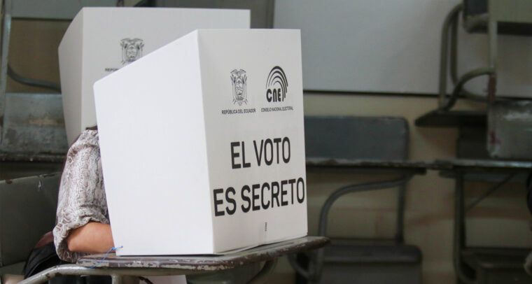 Persona votando detrás de un biombo blanco con la inscripción "El voto es secreto" y logotipos del CNE y la República del Ecuador, en un aula adaptada como recinto electoral con escritorios visibles al fondo. Elecciones