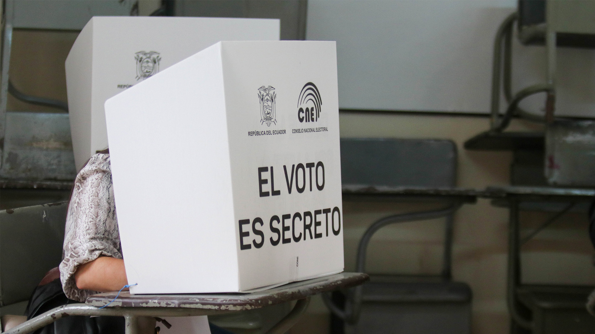 Persona votando detrás de un biombo blanco con la inscripción "El voto es secreto" y logotipos del CNE y la República del Ecuador, en un aula adaptada como recinto electoral con escritorios visibles al fondo. Elecciones