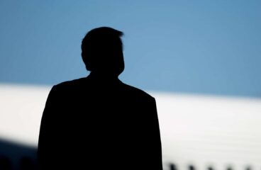 En primer plano, se aprecia la silueta de Donald Trump durante la ceremonia de presentación del Boeing 787-10 en Carolina del Sur, Estados Unidos. Al fondo, se distingue el perfil de un avión blanco bajo un cielo azul en pleno día.