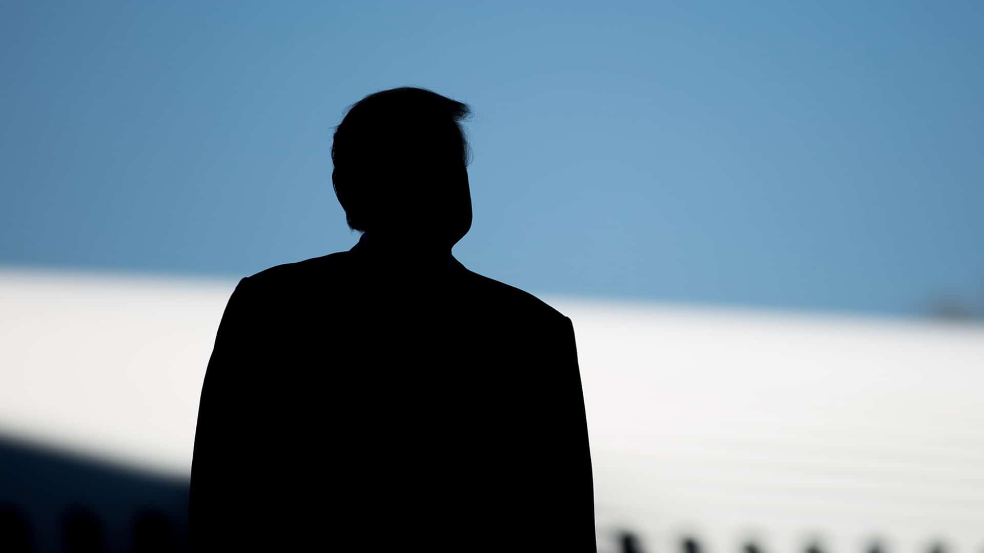 En primer plano, se aprecia la silueta de Donald Trump durante la ceremonia de presentación del Boeing 787-10 en Carolina del Sur, Estados Unidos. Al fondo, se distingue el perfil de un avión blanco bajo un cielo azul en pleno día.