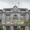 Fachada principal del edificio del Banco de España en Madrid, con arquitectura neoclásica y detalles ornamentales en piedra. Se observan dos farolas en primer plano, árboles a los lados y un cielo nublado. Vehículos en movimiento crean un efecto de desenfoque frente al edificio. Economía