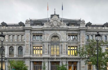 Fachada principal del edificio del Banco de España en Madrid, con arquitectura neoclásica y detalles ornamentales en piedra. Se observan dos farolas en primer plano, árboles a los lados y un cielo nublado. Vehículos en movimiento crean un efecto de desenfoque frente al edificio. Economía