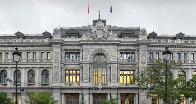 Fachada principal del edificio del Banco de España en Madrid, con arquitectura neoclásica y detalles ornamentales en piedra. Se observan dos farolas en primer plano, árboles a los lados y un cielo nublado. Vehículos en movimiento crean un efecto de desenfoque frente al edificio. Economía