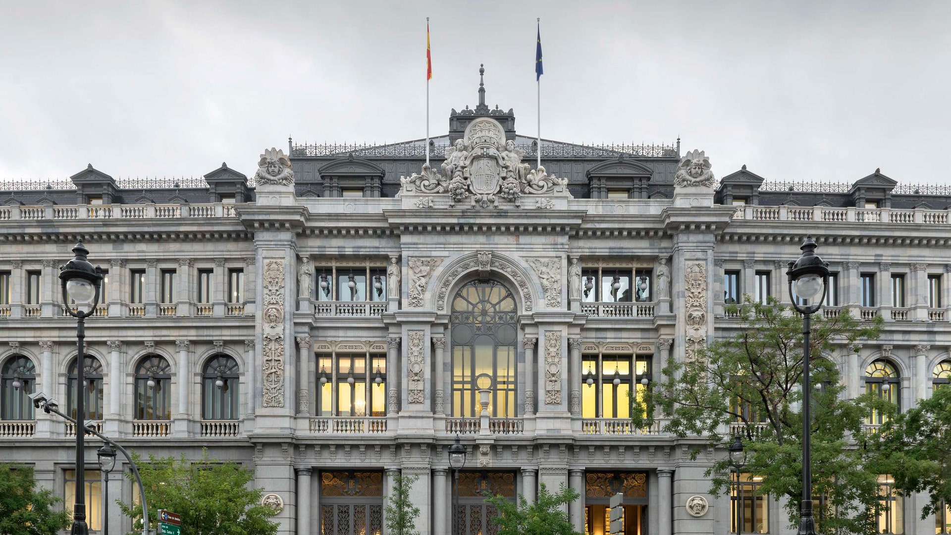 Fachada principal del edificio del Banco de España en Madrid, con arquitectura neoclásica y detalles ornamentales en piedra. Se observan dos farolas en primer plano, árboles a los lados y un cielo nublado. Vehículos en movimiento crean un efecto de desenfoque frente al edificio. Economía