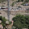 Tres soldados estadounidenses observan el cruce fronterizo de Torkham, situado entre Pakistán y Afganistán, desde una colina, con un paisaje árido y montañoso al fondo. Se observa una carretera con tráfico y peatones cruzando, delimitada por una cerca metálica. Hay árboles y vegetación en primer plano. Afganistán