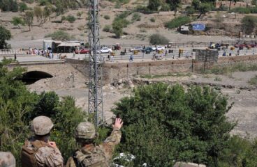 Tres soldados estadounidenses observan el cruce fronterizo de Torkham, situado entre Pakistán y Afganistán, desde una colina, con un paisaje árido y montañoso al fondo. Se observa una carretera con tráfico y peatones cruzando, delimitada por una cerca metálica. Hay árboles y vegetación en primer plano. Afganistán