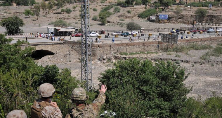 Tres soldados estadounidenses observan el cruce fronterizo de Torkham, situado entre Pakistán y Afganistán, desde una colina, con un paisaje árido y montañoso al fondo. Se observa una carretera con tráfico y peatones cruzando, delimitada por una cerca metálica. Hay árboles y vegetación en primer plano. Afganistán