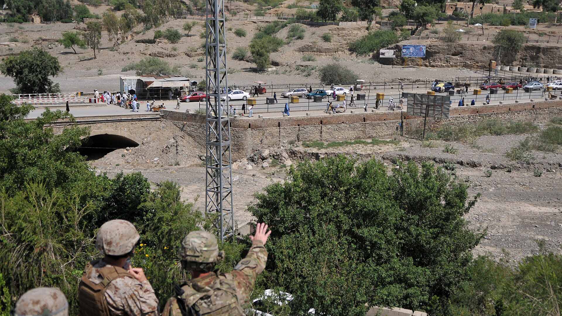 Tres soldados estadounidenses observan el cruce fronterizo de Torkham, situado entre Pakistán y Afganistán, desde una colina, con un paisaje árido y montañoso al fondo. Se observa una carretera con tráfico y peatones cruzando, delimitada por una cerca metálica. Hay árboles y vegetación en primer plano. Afganistán