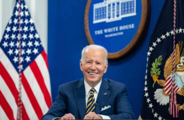 Joe Biden (sentado, con un traje azul, camisa blanca y corbata de rayas azules y amarillas) durante una llamada virtual con simpatizantes para conmemorar el primer aniversario de su administración, el miércoles 19 de enero de 2022, en el Auditorio South Court del Edificio de Oficinas Ejecutivas Eisenhower de la Casa Blanca. Detrás, la bandera de EEUU y el logo oficial de la Casa Blanca