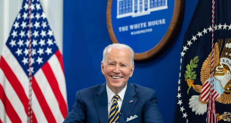 Joe Biden (sentado, con un traje azul, camisa blanca y corbata de rayas azules y amarillas) durante una llamada virtual con simpatizantes para conmemorar el primer aniversario de su administración, el miércoles 19 de enero de 2022, en el Auditorio South Court del Edificio de Oficinas Ejecutivas Eisenhower de la Casa Blanca. Detrás, la bandera de EEUU y el logo oficial de la Casa Blanca