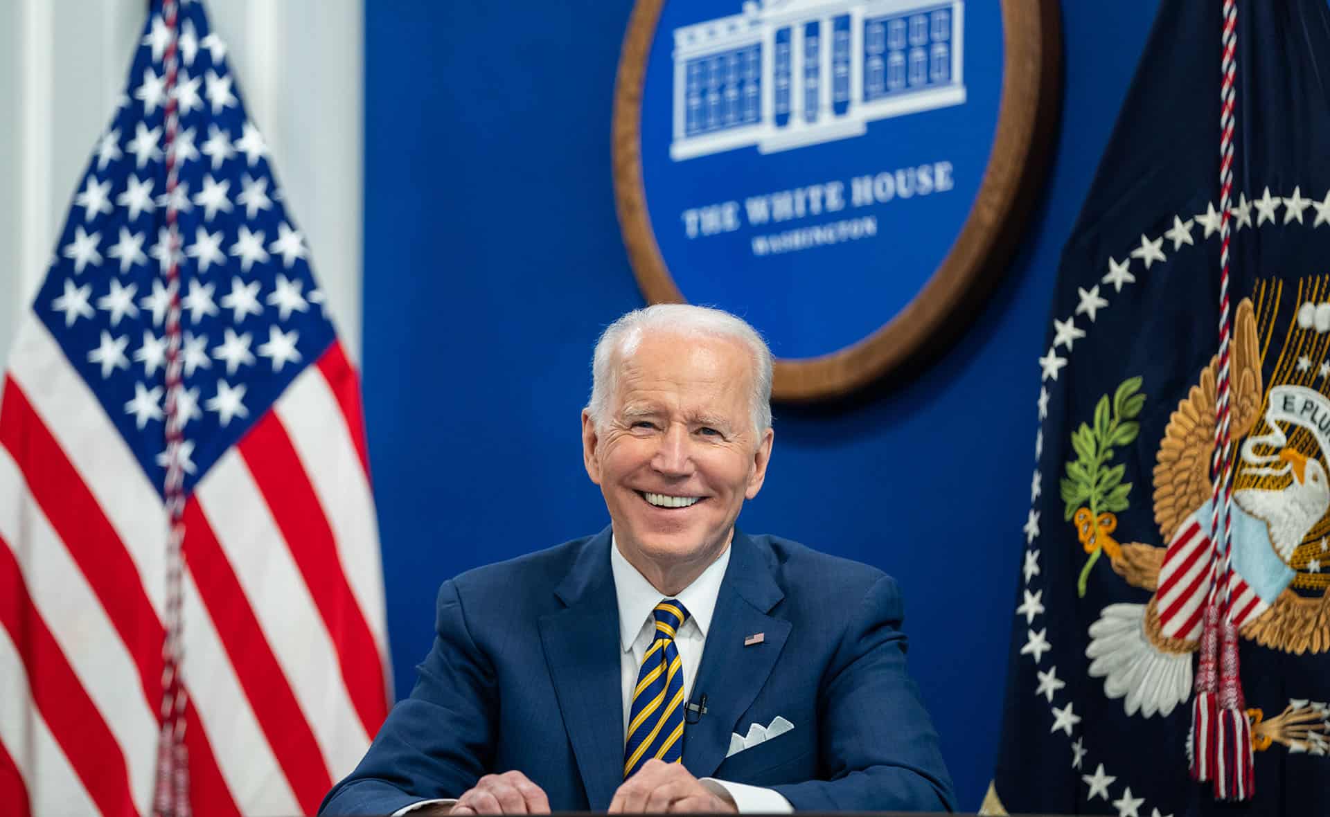 Joe Biden (sentado, con un traje azul, camisa blanca y corbata de rayas azules y amarillas) durante una llamada virtual con simpatizantes para conmemorar el primer aniversario de su administración, el miércoles 19 de enero de 2022, en el Auditorio South Court del Edificio de Oficinas Ejecutivas Eisenhower de la Casa Blanca. Detrás, la bandera de EEUU y el logo oficial de la Casa Blanca