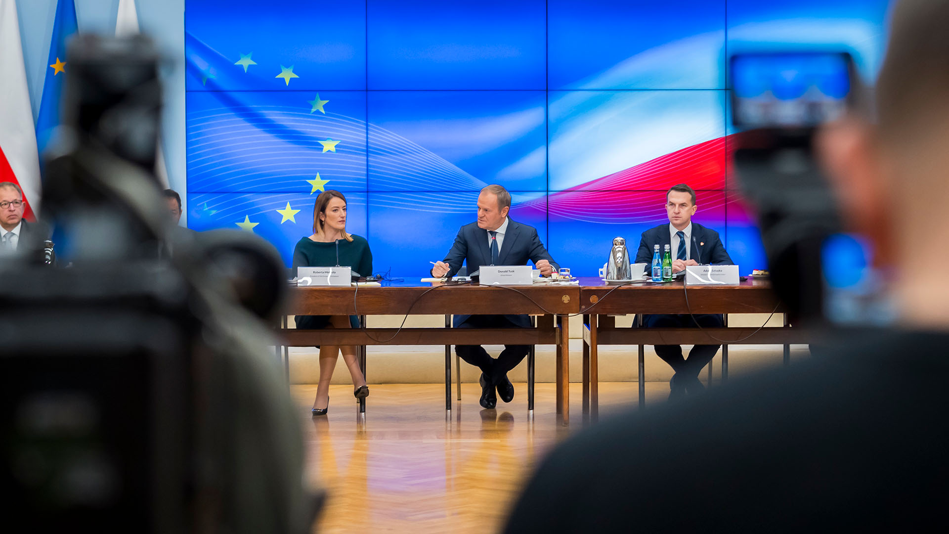 Reunión oficial de la Conferencia de Presidentes del Parlamento Europeo con el primer ministro polaco Donald Tusk y representantes del gobierno de Polonia. En el fondo, se observa una pantalla con la bandera de la Unión Europea y la bandera de Polonia, mientras los participantes dialogan en torno a una mesa en un entorno institucional. Presidencia