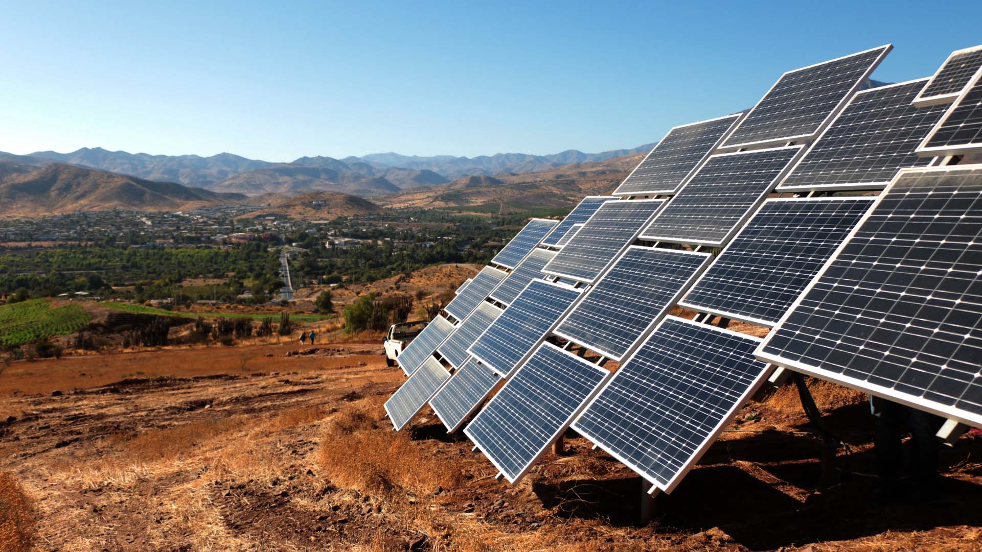 A series of solar panels installed on a hillside near the city of Ovalle (Chile) set against a rural landscape with distant mountains under the clear blue sky of Monte Patria. Colombia and Chile