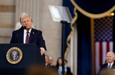 Donald Trump pronuncia un discurso durante su ceremonia de investidura como presidente de los Estados Unidos, en Washington, el 20 de enero de 2025. Está de pie detrás de un atril con el escudo presidencial. Lleva un traje azul marino, una camisa blanca y una corbata roja oscura. Al fondo, se aprecia una bandera de los Estados Unidos. Asimismo, se percibe, de forma desenfocada, la figura de Joe Biden, expresidente de los Estados Unidos. Foto: The Trump White House (Wikimedia Commons/Dominio público). Trump