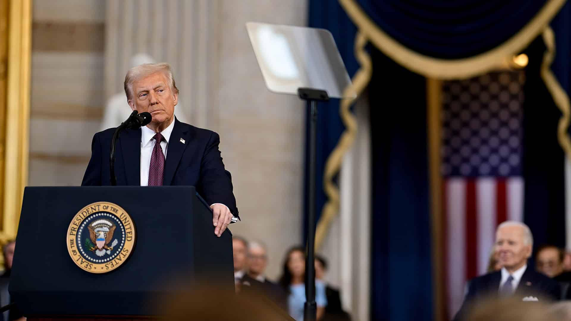 Donald Trump pronuncia un discurso durante su ceremonia de investidura como presidente de los Estados Unidos, en Washington, el 20 de enero de 2025. Está de pie detrás de un atril con el escudo presidencial. Lleva un traje azul marino, una camisa blanca y una corbata roja oscura. Al fondo, se aprecia una bandera de los Estados Unidos. Asimismo, se percibe, de forma desenfocada, la figura de Joe Biden, expresidente de los Estados Unidos. Foto: The Trump White House (Wikimedia Commons/Dominio público). Trump