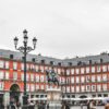 Vista de la Plaza Mayor de Madrid (España) en un día nublado. En el centro resalta la estatua ecuestre del Rey Felipe III, rodeada de edificios de fachada roja, tejados de pizarra gris y balcones alineados. Los soportales de la planta baja albergan cafés y tiendas, mientras se aprecia a varias personas caminando. En primer plano, destaca una farola negra ornamentada. Madrid