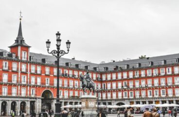 View of Plaza Mayor in Madrid (Spain) on a cloudy day. At the centre, the equestrian statue of King Philip III stands out, surrounded by red-facade buildings, grey slate roofs, and aligned balconies. The arcades on the ground floor host cafés and shops, with several people visible walking around. In the foreground, an ornate black lamppost is prominent. Madrid