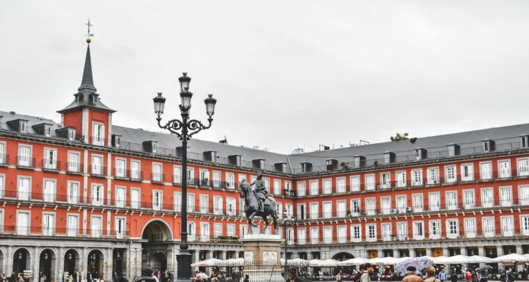 Vista de la Plaza Mayor de Madrid (España) en un día nublado. En el centro resalta la estatua ecuestre del Rey Felipe III, rodeada de edificios de fachada roja, tejados de pizarra gris y balcones alineados. Los soportales de la planta baja albergan cafés y tiendas, mientras se aprecia a varias personas caminando. En primer plano, destaca una farola negra ornamentada. Madrid