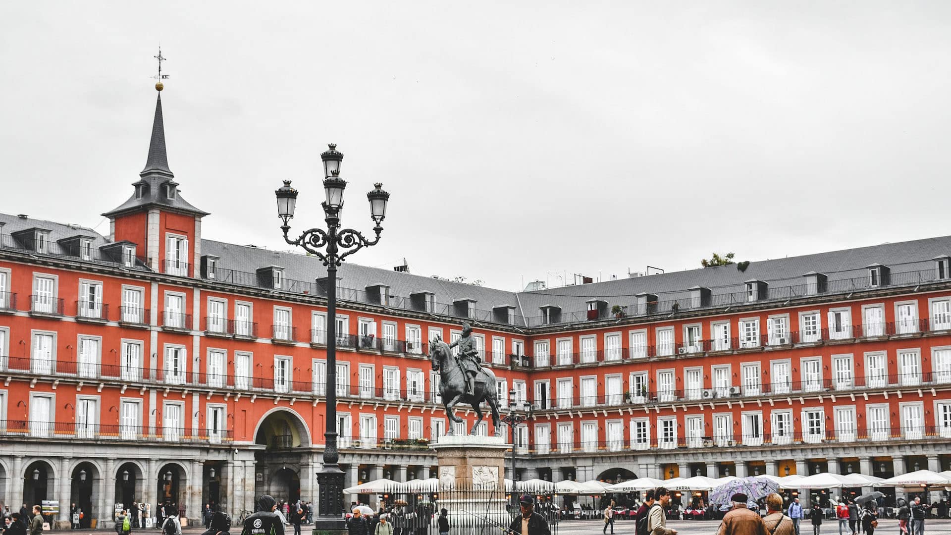 View of Plaza Mayor in Madrid (Spain) on a cloudy day. At the centre, the equestrian statue of King Philip III stands out, surrounded by red-facade buildings, grey slate roofs, and aligned balconies. The arcades on the ground floor host cafés and shops, with several people visible walking around. In the foreground, an ornate black lamppost is prominent. Madrid