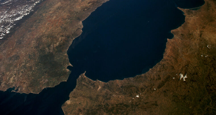 Vista aérea diurna del Estrecho de Gibraltar, separando España (norte) y Marruecos (sur). Se observan la Sierra Nevada en España, la costa montañosa de Marruecos, el río Guadalquivir y las ciudades de Gibraltar y Ceuta bajo cielos despejados. Ceuta y Melilla