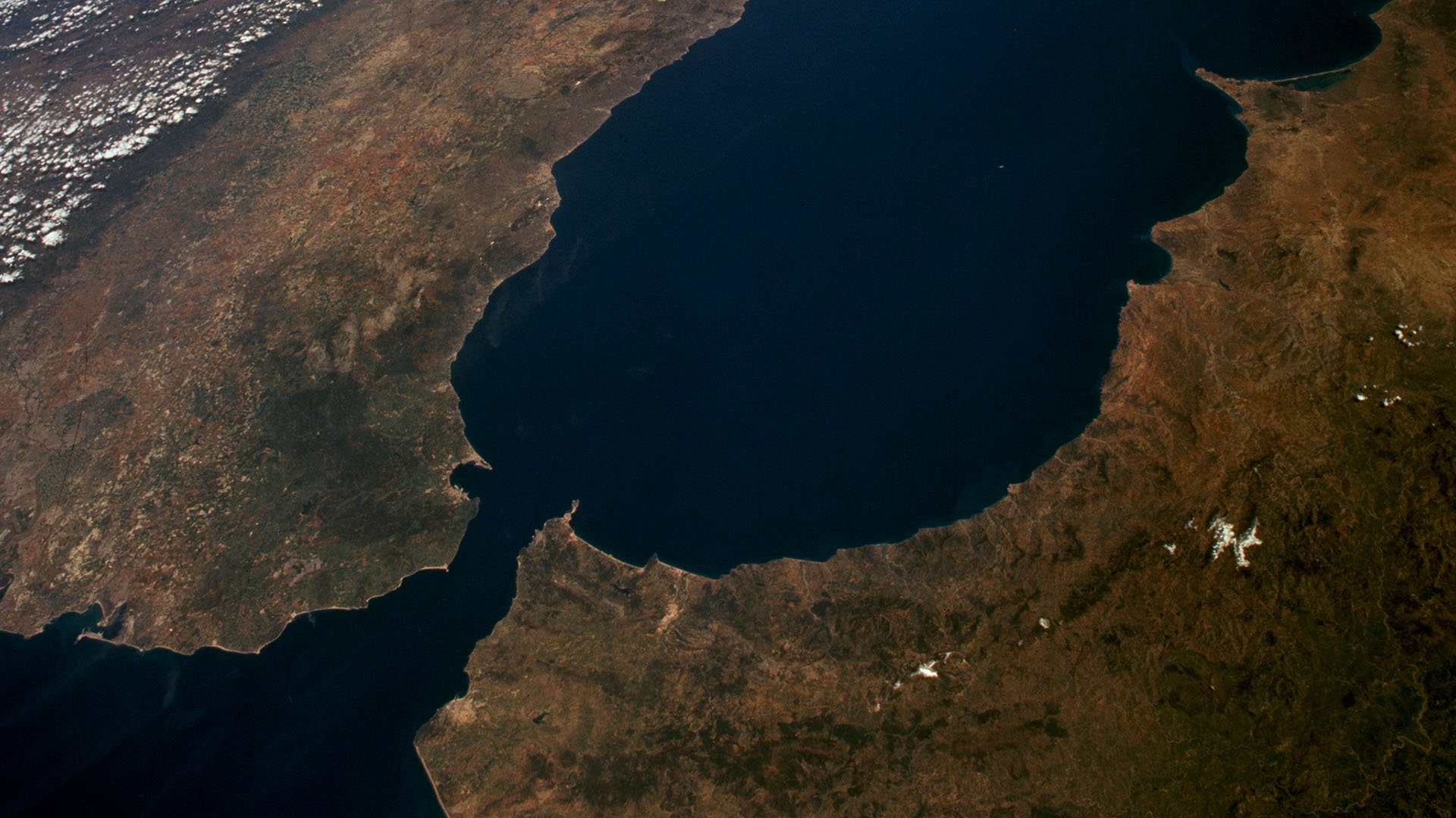 Daytime aerial view of the Strait of Gibraltar, separating Spain (north) and Morocco (south). Visible features include the Sierra Nevada in Spain, the mountainous coastline of Morocco, the River Guadalquivir, and the cities of Gibraltar and Ceuta under clear skies. Ceuta and Melilla