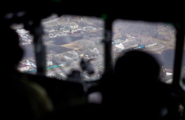 Vista aérea de una zona del este de Ucrania a través de la ventana de una aeronave en un día nublado. En primer plano, se distinguen las siluetas oscuras de dos soldados ucranianos que observan el terreno desde el interior de la cabina. En el exterior, el paisaje muestra casas dispersas con techos de chapa, calles angostas y terrenos agrícolas. Ucrania