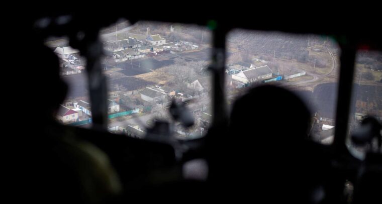 Vista aérea de una zona del este de Ucrania a través de la ventana de una aeronave en un día nublado. En primer plano, se distinguen las siluetas oscuras de dos soldados ucranianos que observan el terreno desde el interior de la cabina. En el exterior, el paisaje muestra casas dispersas con techos de chapa, calles angostas y terrenos agrícolas. Ucrania