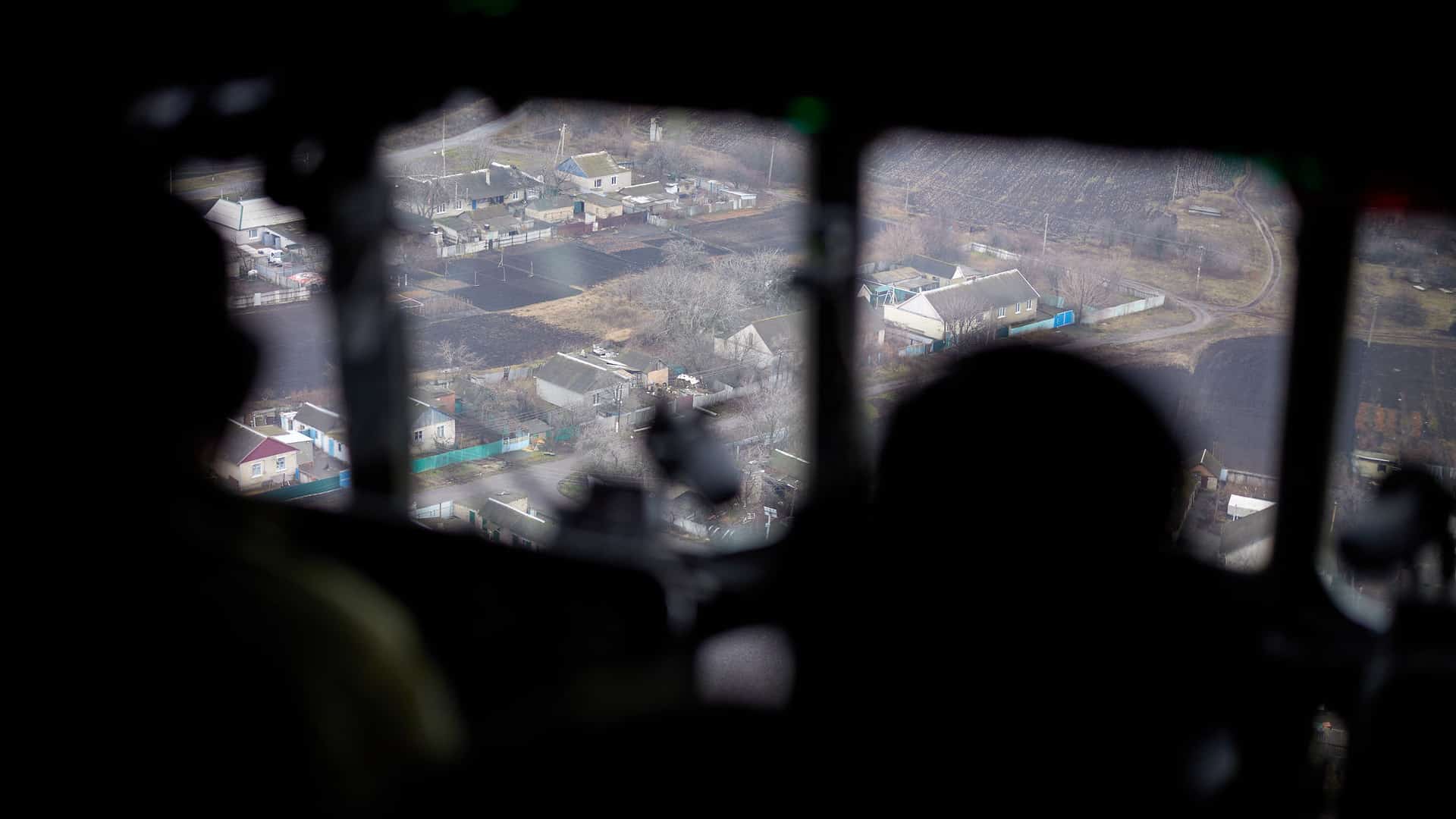 Vista aérea de una zona del este de Ucrania a través de la ventana de una aeronave en un día nublado. En primer plano, se distinguen las siluetas oscuras de dos soldados ucranianos que observan el terreno desde el interior de la cabina. En el exterior, el paisaje muestra casas dispersas con techos de chapa, calles angostas y terrenos agrícolas. Ucrania