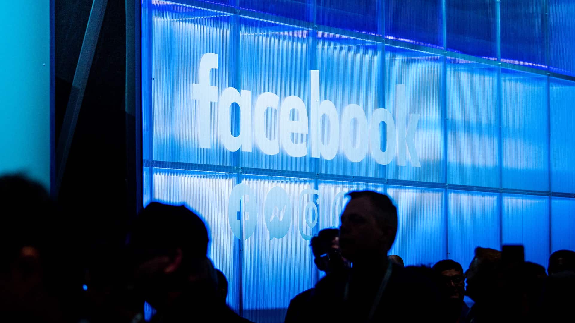 Logotipo iluminado de Facebook sobre una pared azul translúcida en la conferencia Facebook F8 2019. Debajo del logotipo se aprecian también los íconos de Messenger e Instagram. En primer plano, siluetas de asistentes contrastan con la luz azul. Meta