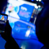 Close-up of a hand holding a mobile phone displaying a live broadcast of Roberta Metsola (seated in a studio, wearing a light-coloured suit and speaking into a microphone), President of the European Parliament. The blurred background reveals the shadow of a person and the same studio, illuminated by blue lights. digital