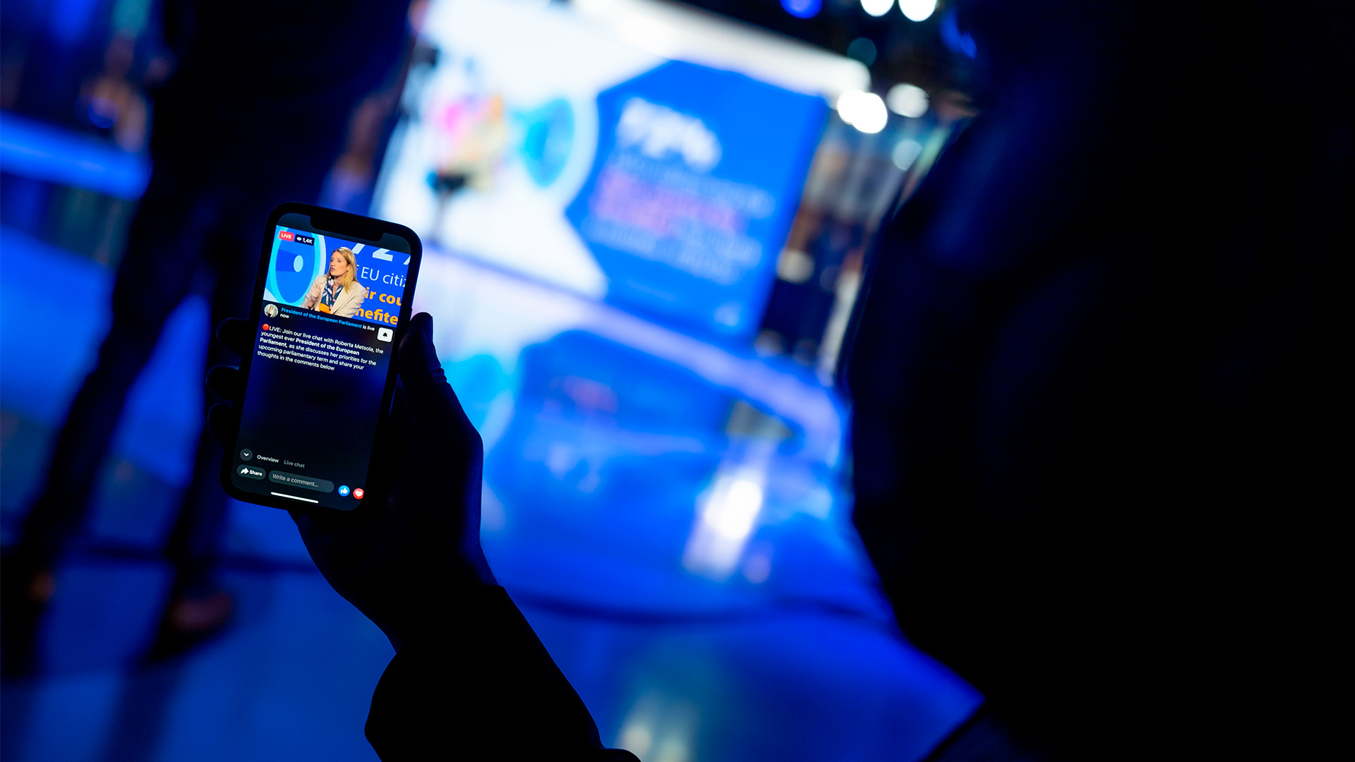 Close-up of a hand holding a mobile phone displaying a live broadcast of Roberta Metsola (seated in a studio, wearing a light-coloured suit and speaking into a microphone), President of the European Parliament. The blurred background reveals the shadow of a person and the same studio, illuminated by blue lights. digital