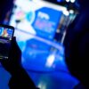 A hand holding a mobile phone showing a live broadcast on the Facebook page of the European Parliament (EP) of Roberta Metsola, EP President, sitting in a studio, wearing a cream-coloured suit, speaking into a microphone. The blurred background reveals the shadow of a person in the same studio, illuminated by blue lights