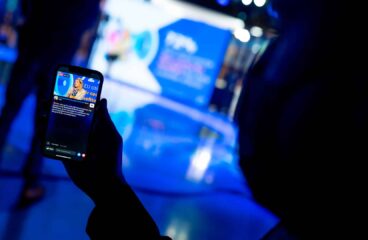 A hand holding a mobile phone showing a live broadcast on the Facebook page of the European Parliament (EP) of Roberta Metsola, EP President, sitting in a studio, wearing a cream-coloured suit, speaking into a microphone. The blurred background reveals the shadow of a person in the same studio, illuminated by blue lights