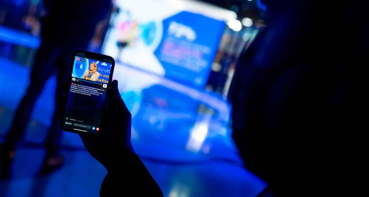 A hand holding a mobile phone showing a live broadcast on the Facebook page of the European Parliament (EP) of Roberta Metsola, EP President, sitting in a studio, wearing a cream-coloured suit, speaking into a microphone. The blurred background reveals the shadow of a person in the same studio, illuminated by blue lights