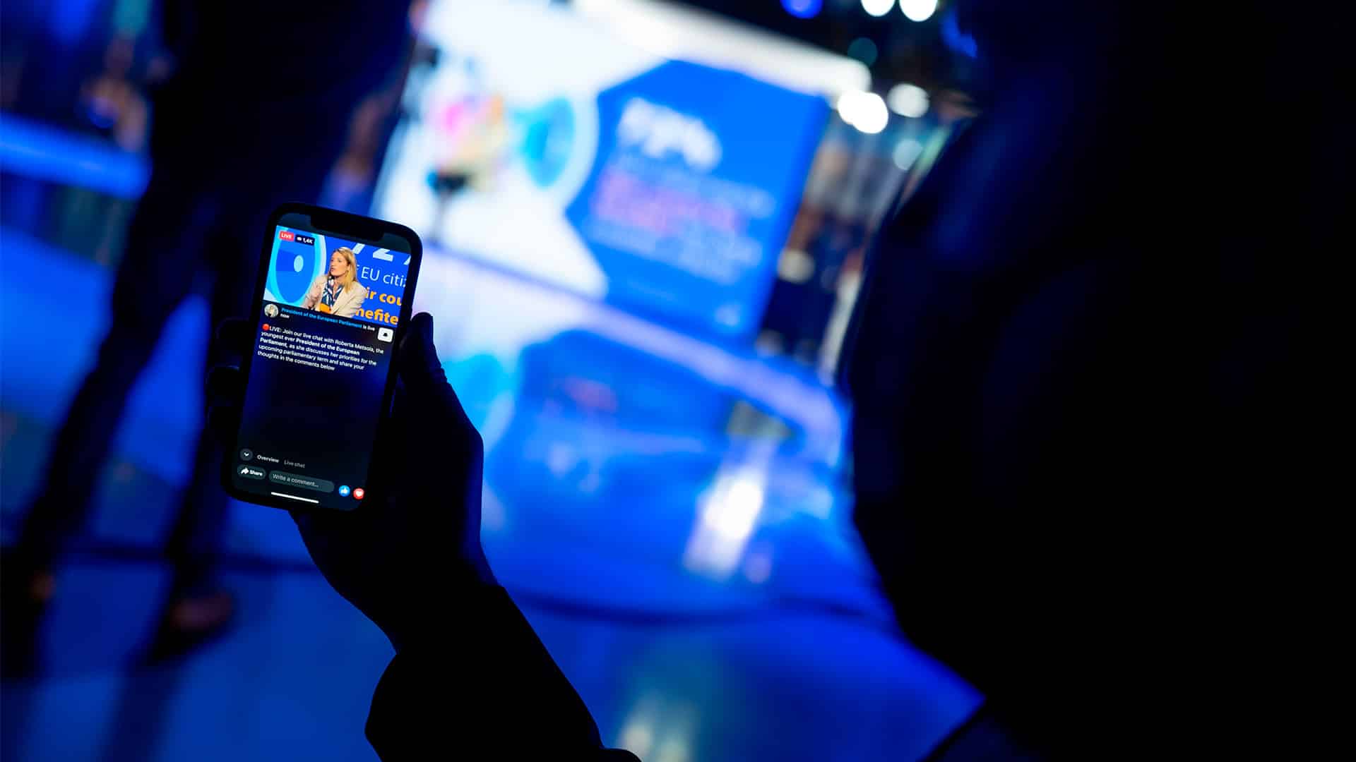 A hand holding a mobile phone showing a live broadcast on the Facebook page of the European Parliament (EP) of Roberta Metsola, EP President, sitting in a studio, wearing a cream-coloured suit, speaking into a microphone. The blurred background reveals the shadow of a person in the same studio, illuminated by blue lights