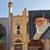 Detalle de la fachada y entrada de la Gran Mezquita de Isfahán (Irán), con su característico arco decorado con azulejos azules y minaretes ornamentados. A la derecha, un gran retrato de Alí Jamenei, líder supremo del país, cuelga sobre un edificio de ladrillo con arcos y motivos geométricos