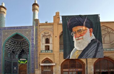 Detalle de la fachada y entrada de la Gran Mezquita de Isfahán (Irán), con su característico arco decorado con azulejos azules y minaretes ornamentados. A la derecha, un gran retrato de Alí Jamenei, líder supremo del país, cuelga sobre un edificio de ladrillo con arcos y motivos geométricos